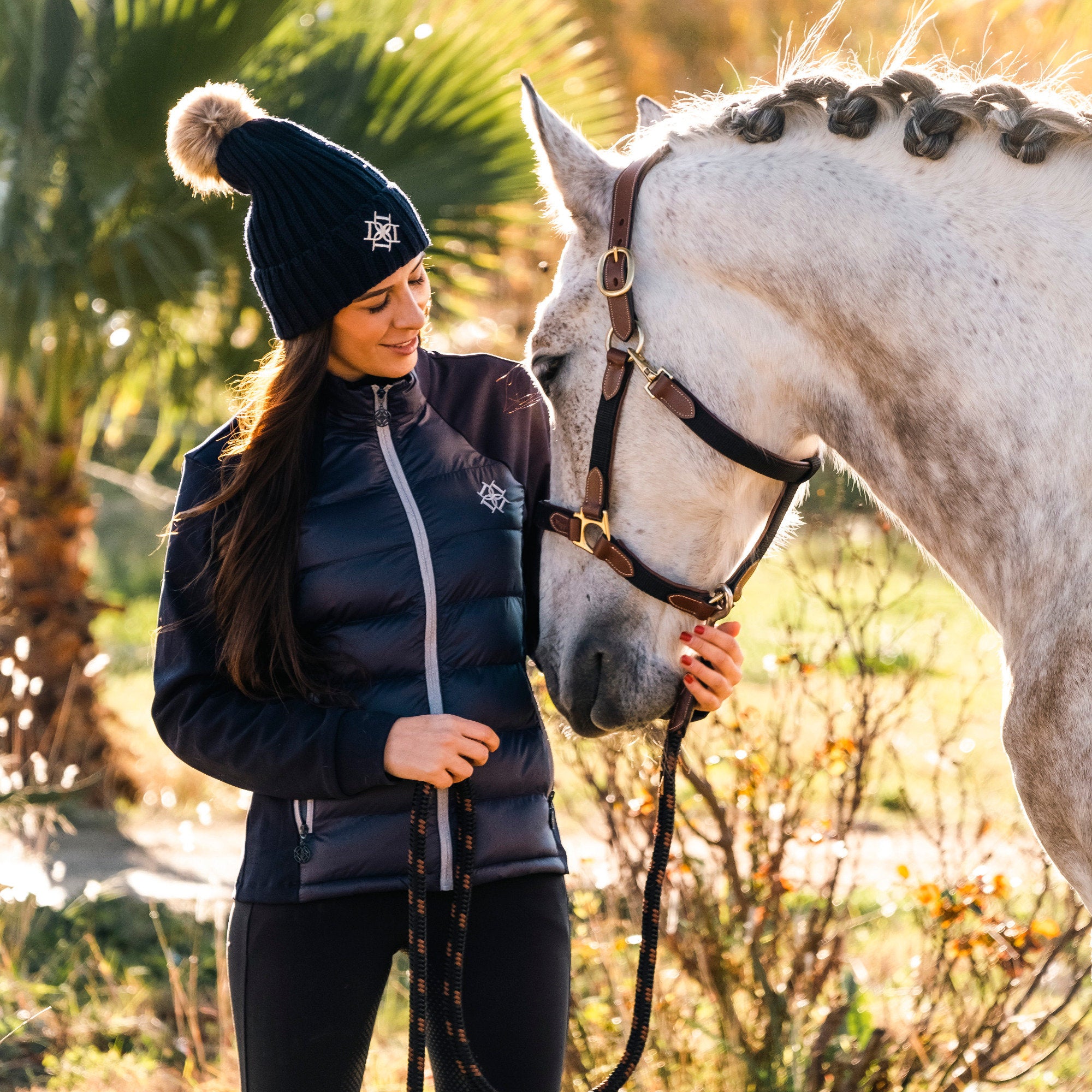 Dapple rider wearing  Hybrid Jacket and Bobble Hat with white horse wearing head collar and lead